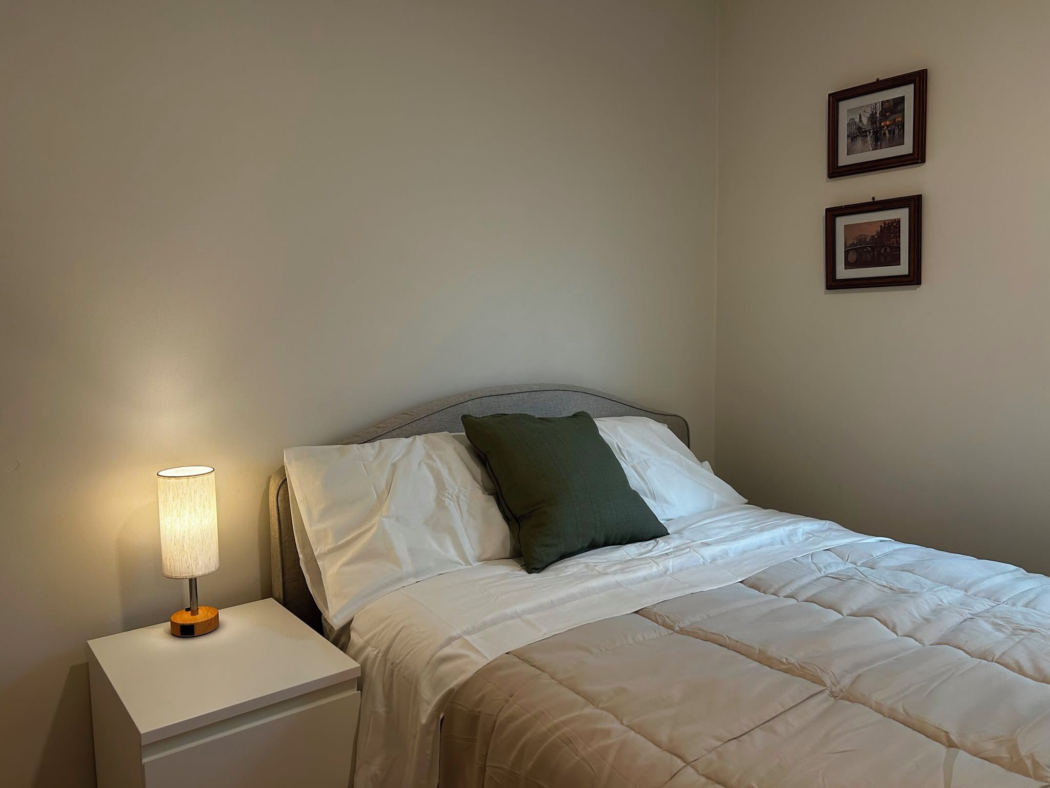 A single bed with white bedding and a green pillow, a bedside table with a lamp, and two framed pictures on the wall.