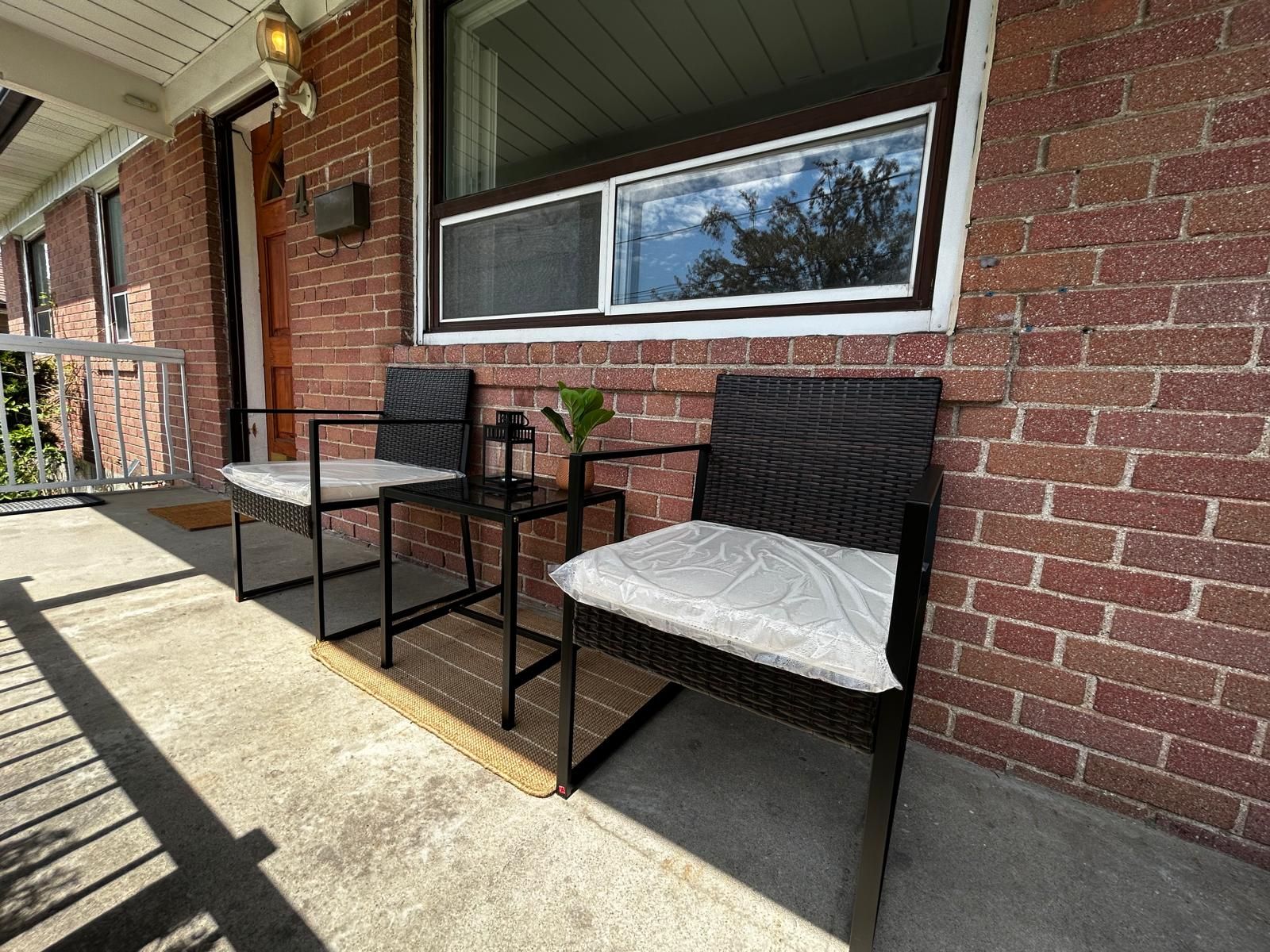 A patio with two wicker chairs and a small table, a brick wall, and a window overlooking a tree.