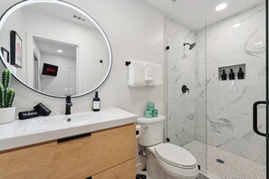 Un bagno moderno con uno specchio rotondo, un lavandino bianco, un armadietto in legno, il water e una cabina doccia in vetro.