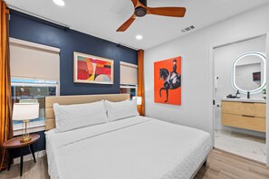 Una camera d'albergo moderna con un letto grande, una ventola a soffitto, un comodino con una lampada, una cassettiera con uno specchio e opere d'arte alle pareti.