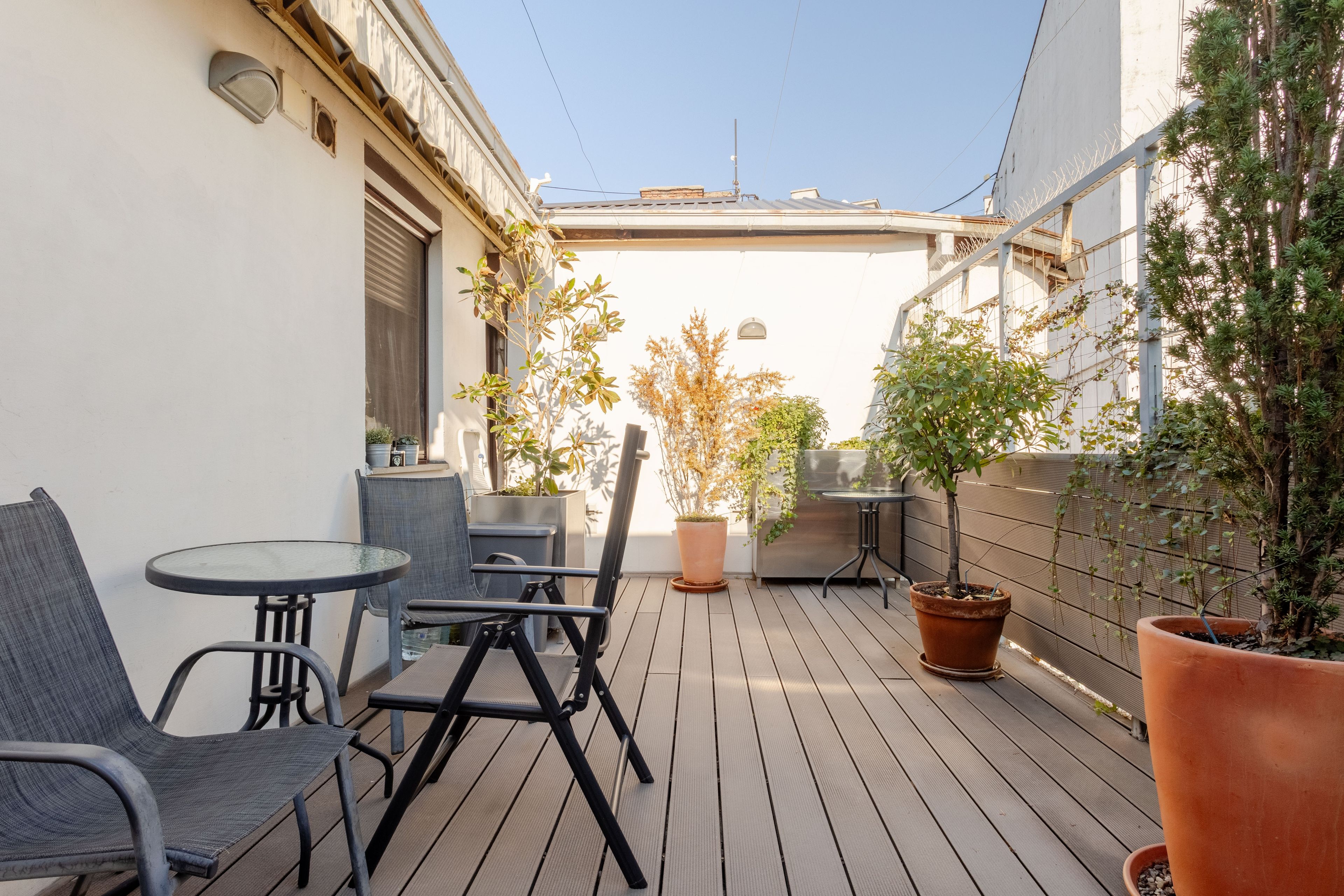 Una terraza de madera con mobiliario exterior, plantas en macetas y cielo despejado.