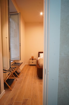 Een smalle gang met een houten vloer, een bed met een wit hoofdeinde, een nachtkastje met een lamp en een raam met uitzicht.
