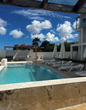 A swimming pool with lounge chairs and umbrellas, surrounded by a building with a covered patio area. - Vrbo Property (Punta Cana)