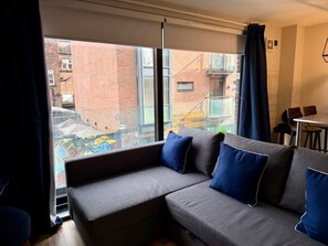 A modern living room with a grey sofa, blue cushions, and a view of a brick building through large windows. - Peaceful 1-bedroom apartment in the heart of Manchester (Manchester)