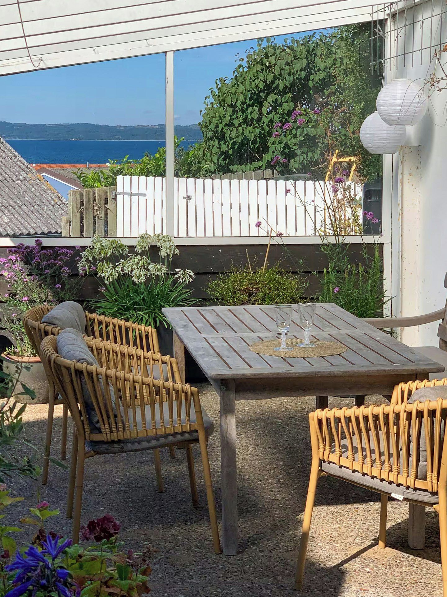 An outdoor patio with a table and chairs, surrounded by plants and a view of the sea.