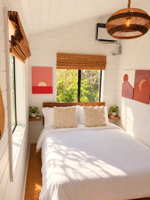 A bedroom with a bed, a window with a view of greenery, a wall-mounted artwork, and a hanging light fixture.