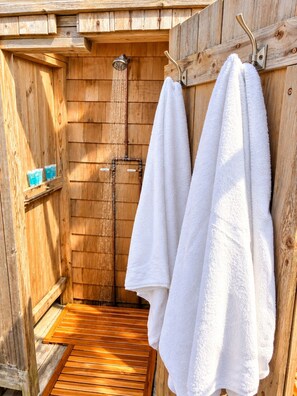 Box doccia in legno con soffione a pioggia, un portasciugamani con due asciugamani bianchi e uno scaffale con articoli da bagno blu.