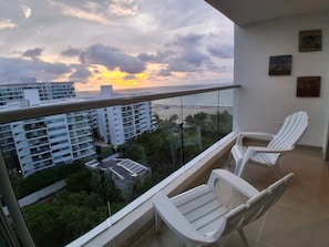 A balcony with two white chairs, a glass railing, and a view of buildings and a sunset. - Apartment at Hotel Sonesta - Morros with sea view and direct beach access | Pool (Cartagena de Indias)