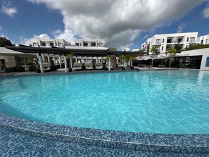 A swimming pool with a tiled edge, surrounded by modern buildings and covered seating areas. - Vista Cana Panorama Lake | 1-Bedroom with King Bed | 1.5 Bathrooms | Pool & Gym (Punta Cana)