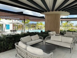 A covered outdoor seating area with wicker furniture, a pool, and a view of the resort. - Vista Cana Panorama Lake | 1-Bedroom with King Bed | 1.5 Bathrooms | Pool & Gym (Punta Cana)