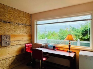 Una habitación con escritorio, una silla roja, una lámpara y una ventana con vistas a zonas verdes.