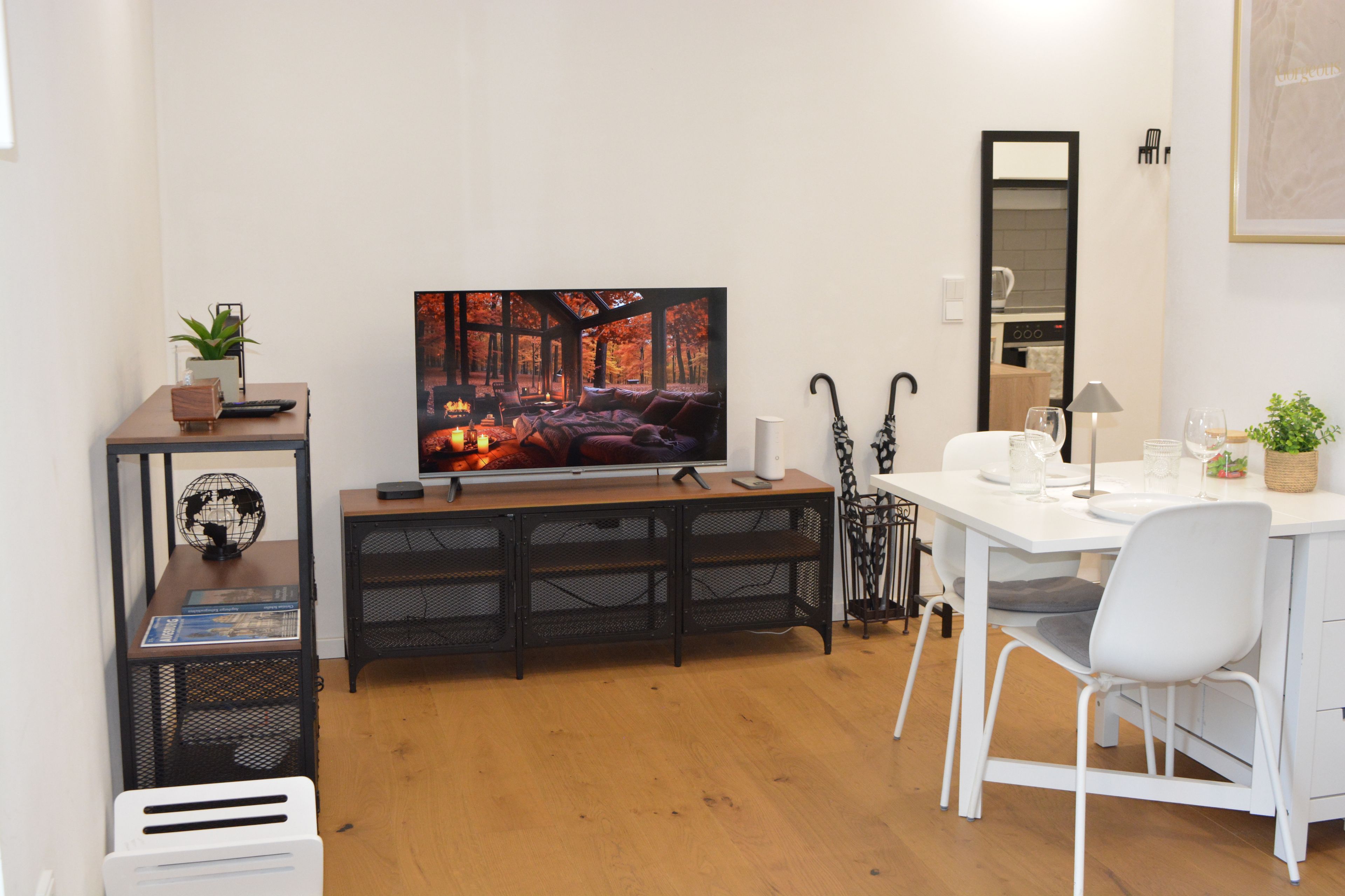 A modern living room with a large flat-screen TV, wooden flooring, a white dining table with chairs, a small bookshelf, and a potted plant.