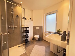 Et moderne badeværelse med et glasafskærmet brusebad, et hvidt toilet, en stor vask med et vindue over, og et håndklædestativ med håndklæder.