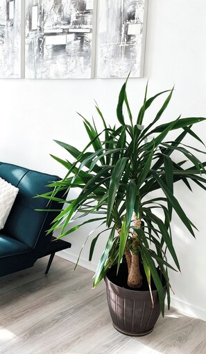 A potted plant in a dark pot on a wooden floor, a blue sofa with a white cushion, and a white wall with abstract artwork. - 2Bedroom Apartment near Disneyland paris,outlet shopping, 2Free Parking, Balcony (Serris)