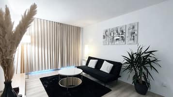 A modern living room with a black sofa, a round coffee table, a large potted plant, and a window with vertical blinds.