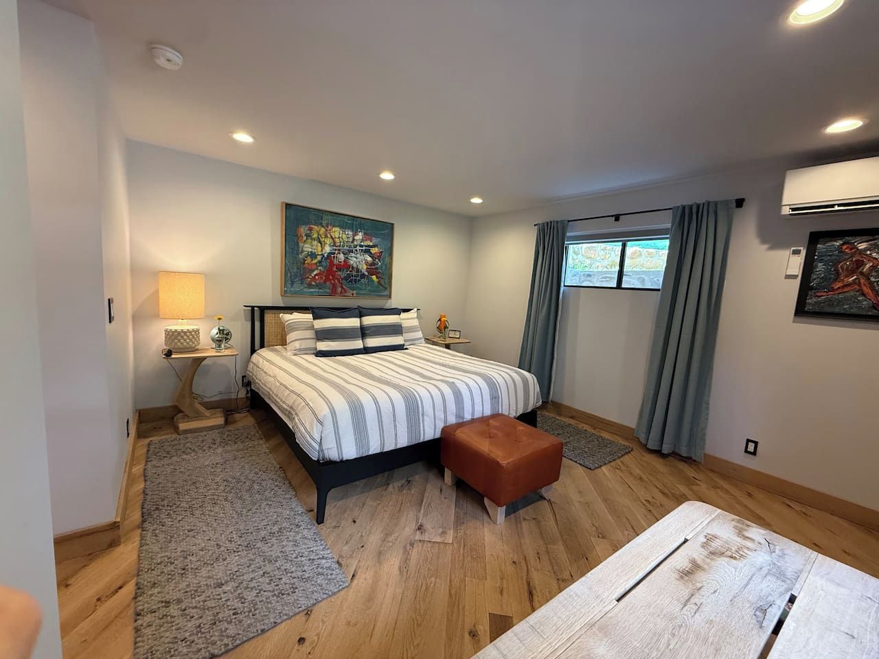 A bedroom with a bed, a nightstand, a lamp, a rug, a wooden bench, and artwork on the walls.