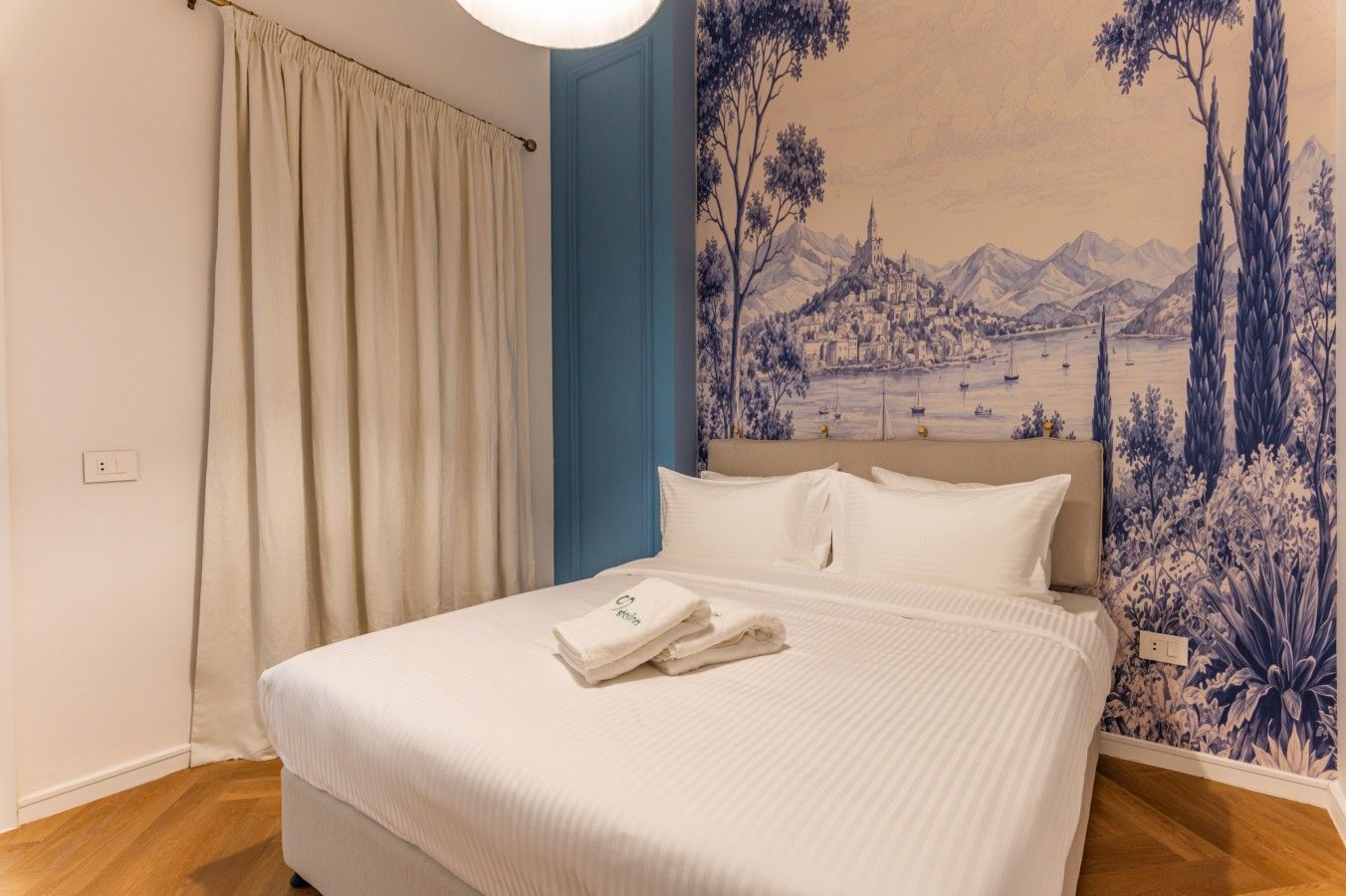 A hotel room with a bed, a curtain, a wall mural, and a wooden floor.