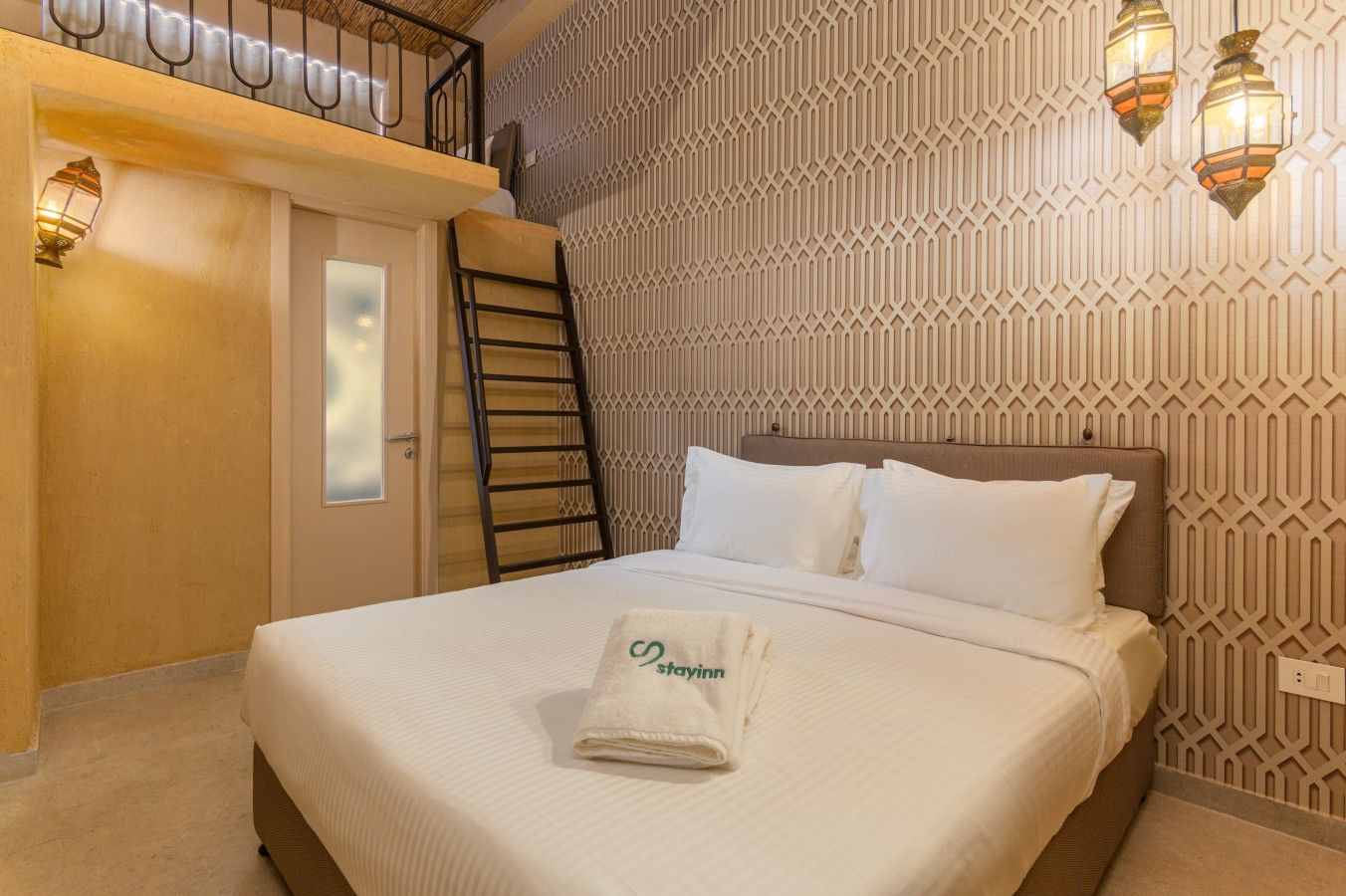 A hotel room with a bed, a ladder, a wall with a geometric pattern, and two wall-mounted lamps.
