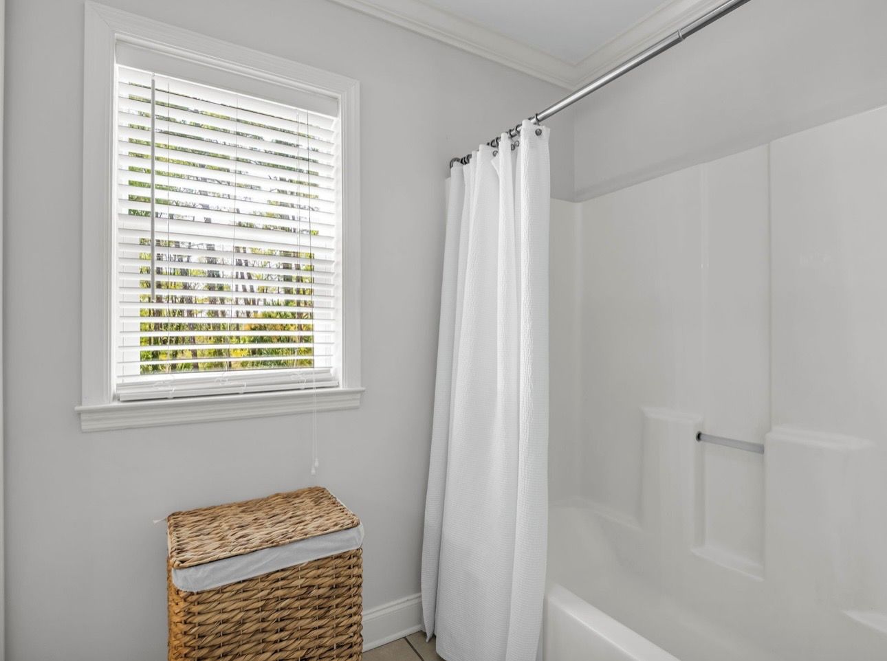 A bathroom with a shower, a window with blinds, and a wicker basket.