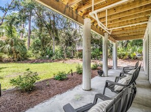 A covered outdoor seating area with chairs and a concrete floor, surrounded by a garden with various plants and trees.