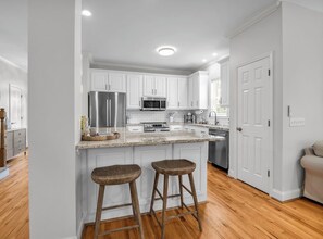 A kitchen with white cabinets, stainless steel appliances, a marble countertop, and wooden stools.