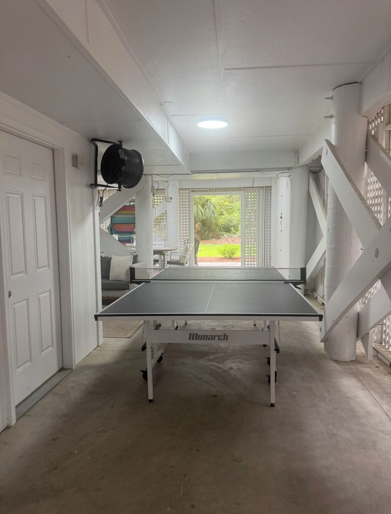 A ping pong table in a room with a white door, a ceiling fan, and a view of a garden through a door.