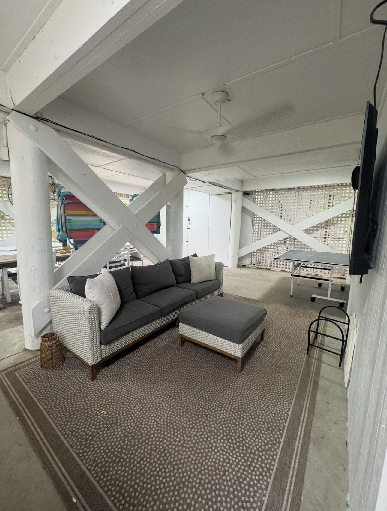 A covered outdoor seating area with a grey sectional sofa, a small ottoman, and a patterned rug.