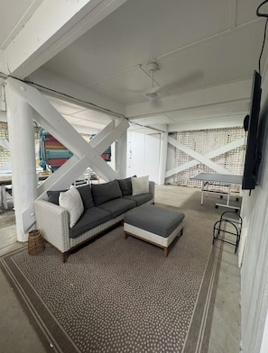 A covered outdoor seating area with a grey sectional sofa, a small ottoman, and a patterned rug.