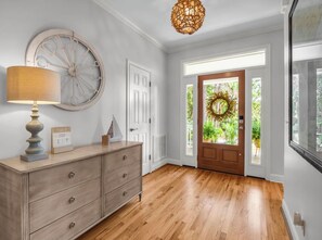 A hallway with a wooden floor, a white door, a wall clock, and a lamp on a dresser.