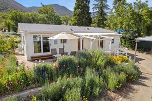 A small, single-story house with a covered patio area, surrounded by a variety of flowering plants and a mountain backdrop. - Peaceful home fully fenced/gated circle driveway and ready for your stay! (Kelseyville)