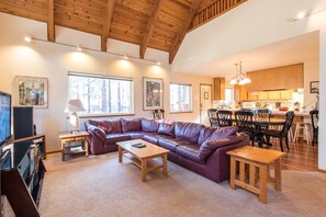 A spacious living room with a large L-shaped sofa, a wooden coffee table, and a television. There is a dining area with a table and chairs, and a kitchen in the background. - Beautiful 5-bedroom chalet in serene South Lake Tahoe perfect for families (South Lake Tahoe)