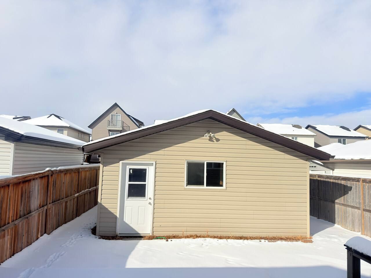 A single-story house with a white door and window, surrounded by a wooden fence, set in a snowy environment.