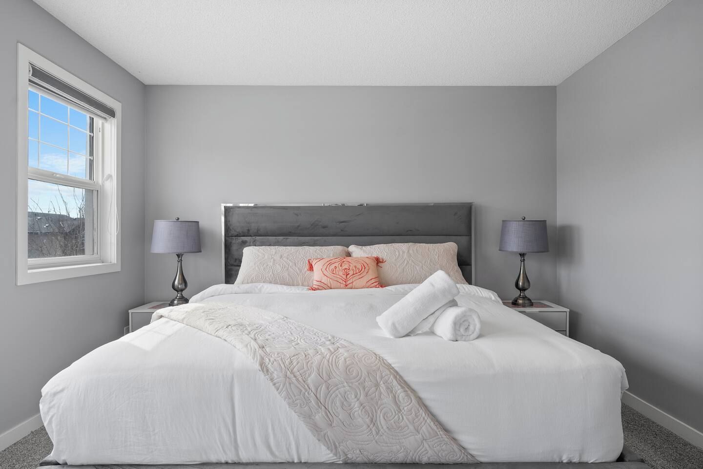 Cama bem arrumada com lençóis brancos, duas luminárias de cabeceira e uma janela com cortinas.