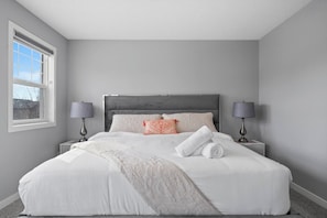 Cama bem arrumada com lençóis brancos, duas luminárias de cabeceira e uma janela com cortinas.