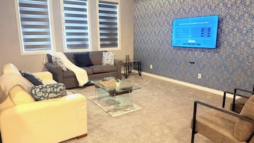 A living room with a sofa, armchair, and coffee table. There are two windows with blinds, a patterned wall, and a flat-screen TV mounted on the wall.