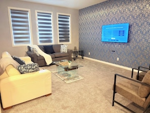 Un salon comprenant un canapé, un fauteuil et une table basse. Il y a deux fenêtres équipées de stores, un mur décoré d’un motif et un téléviseur à écran plat fixé au mur.