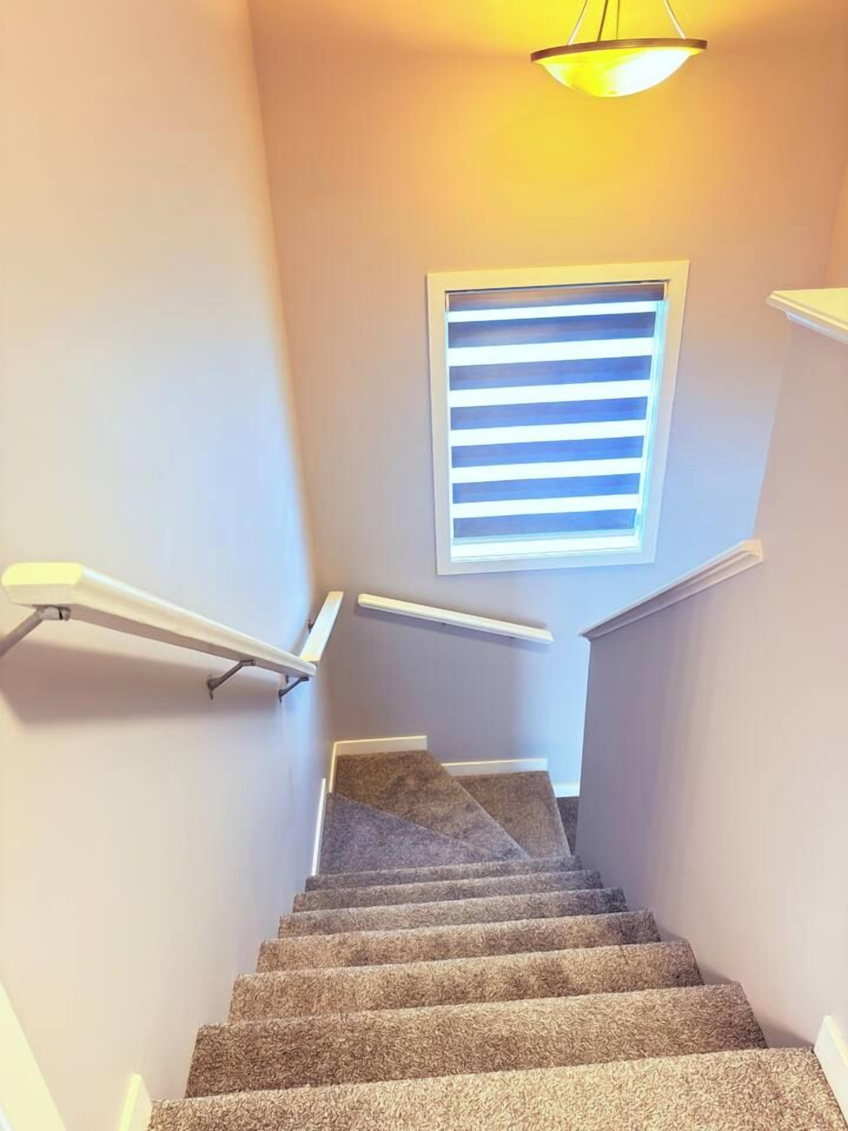 Escadaria coberta de carpete com corrimão, janela com persianas horizontais e luminária no teto.