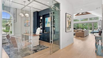 A modern interior with a glass partition separating a dining area from a living space, featuring a kitchen, a sofa, and a large window overlooking greenery.