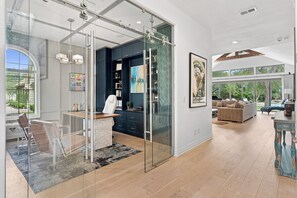 A modern interior with a glass partition separating a dining area from a living space, featuring a kitchen, a sofa, and a large window overlooking greenery. - Austin Chateau Estate: Pool, Hot Tub, Saunas, Cold Plunge, Gym & Theater (Austin)