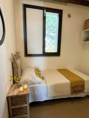 Una cama individual con colchón blanco y manta marrón, una mesita de noche con un jarrón de flores y velas, y una ventana con vista a zonas verdes.