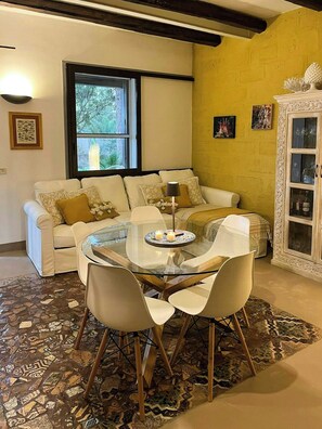 A living room with a glass-top coffee table, white chairs, a sofa with patterned cushions, a cabinet with glass doors, and a window with a view of greenery. - Villa with Outdoor Kitchen and Coastal Garden (Favignana)