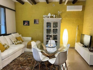 A living room with a white sofa, a glass dining table with chairs, a cabinet with glass doors, and a television on a stand. - Villa with Outdoor Kitchen and Coastal Garden (Favignana)