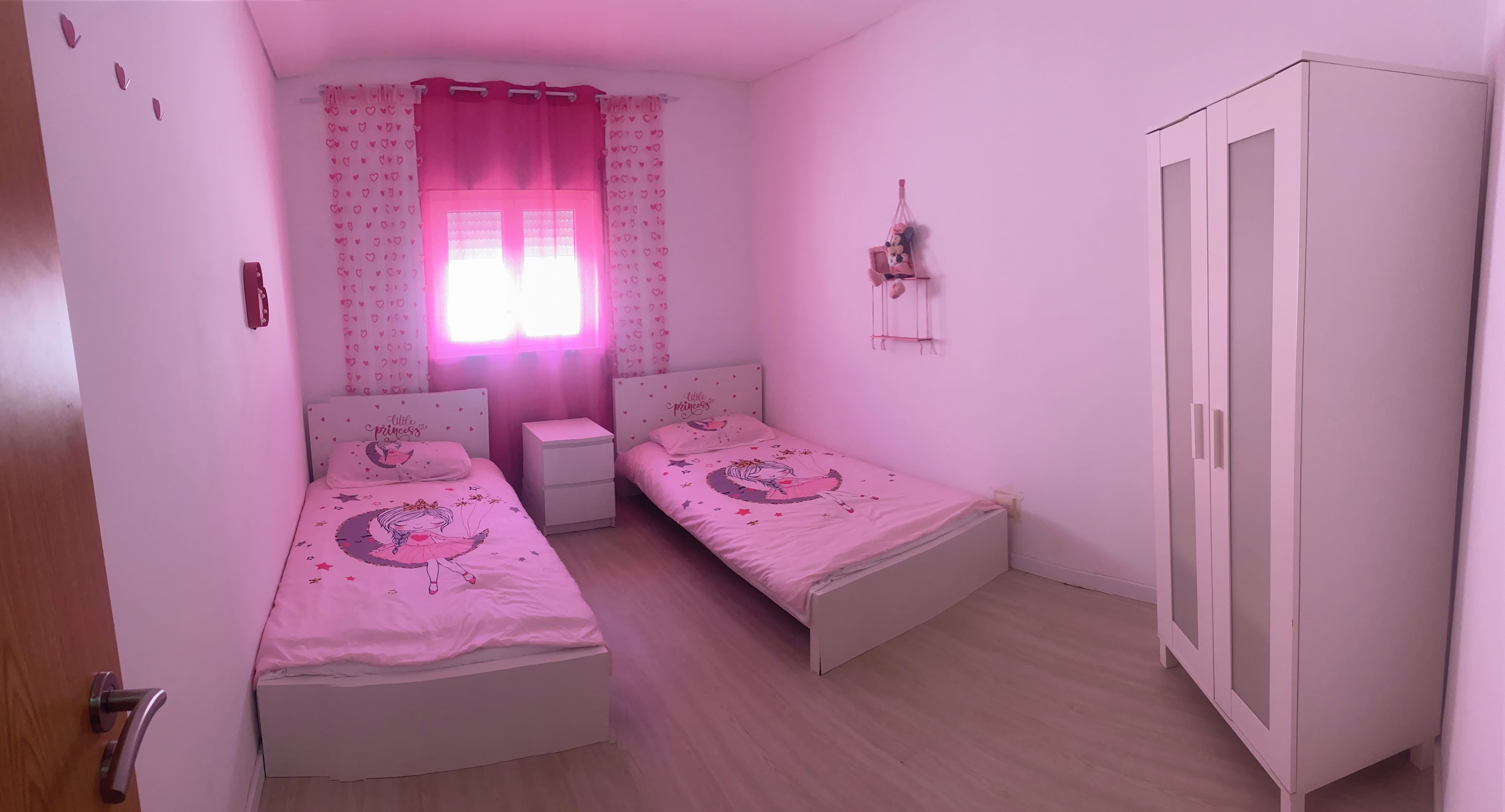 Een slaapkamer met twee eenpersoonsbedden, een kledingkast, een nachtkastje en een raam met hartvormige gordijnen.