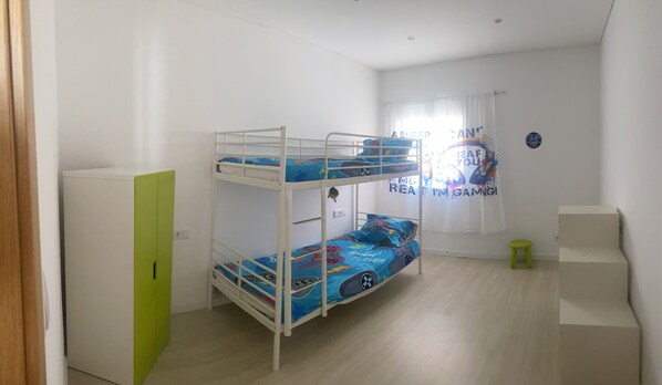 Une chambre avec un lit superposé, un placard vert, un petit tabouret et une fenêtre avec un rideau.