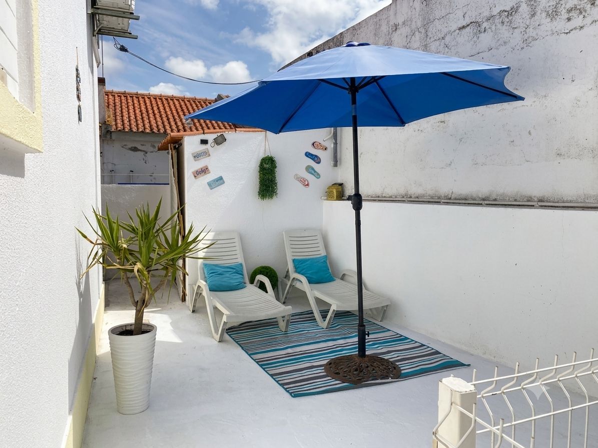 Une petite terrasse extérieure avec un parasol bleu, des chaises longues blanches, une plante en pot et un mur décoratif.