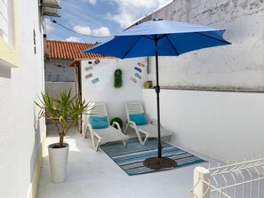 Une petite terrasse extérieure avec un parasol bleu, des chaises longues blanches, une plante en pot et un mur décoratif.