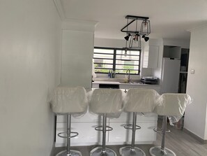Una cocina moderna con armarios blancos, encimera de acero inoxidable y una barra con cuatro taburetes cubiertos de plástico.