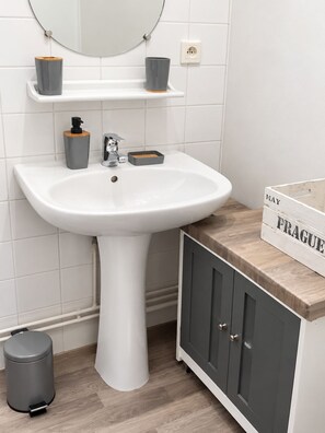 A bathroom with a white pedestal sink, a mirror, a shelf with two containers, a cabinet with drawers, and a trash bin. - Vrbo Property (france)