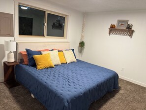 Une chambre à coucher comprenant un lit, une table de chevet, une lampe, une fenêtre, une étagère fixée au mur et une plante suspendue.