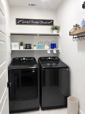 A laundry room with two black washing machines, a shelf with various items, and a sign above. - 3-bedroom house in charming Fort Worth with AC for relaxing stays (Fort Worth)